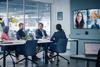 A group of office workers sits together in a meeting, speaking to three people on a large TV screen at the front of the room
