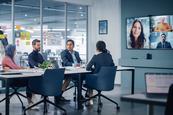 A group of office workers sits together in a meeting, speaking to three people on a large TV screen at the front of the room