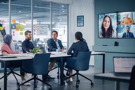A group of office workers sits together in a meeting, speaking to three people on a large TV screen at the front of the room