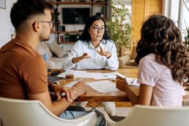 Three people sit around a wooden table in a well-lit room with plants and shelves in the background. A woman in the middle is gesturing with her hands while speaking to the couple across from her, who listen intently