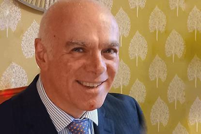 Laurence Howland is a white man in a dark suit and tie, with shaven hair. He's smiling in front of mustard wallpaper with a fan pattern